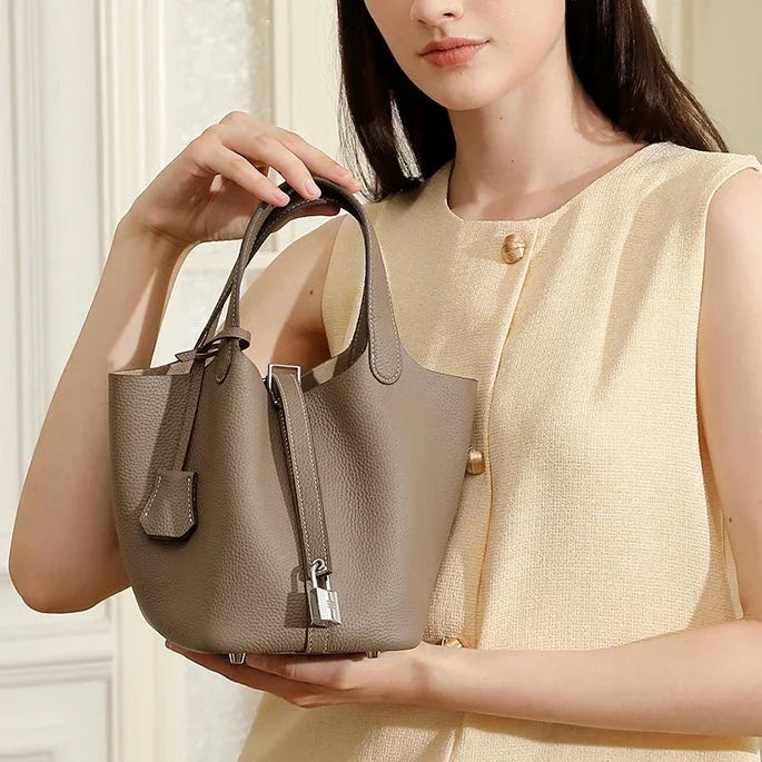 Woman holding taupe leather bucket bag with lock detail, wearing a sleeveless beige top