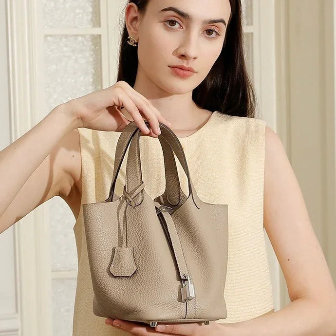 Woman holding a beige leather bucket bag with top handles and padlock detail indoors
