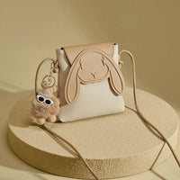 Beige bunny crossbody bag with floppy ears and a fluffy keychain on a textured pedestal
