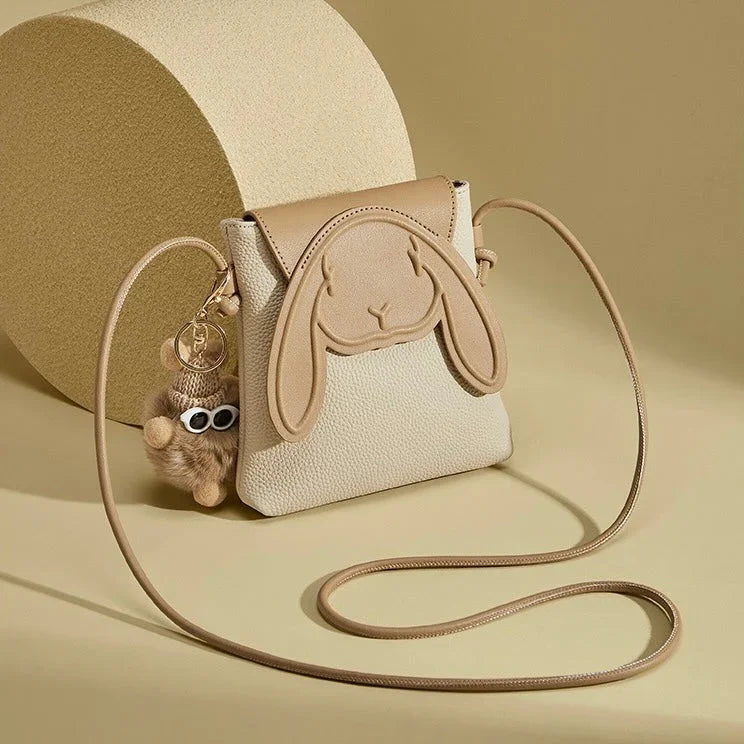 Beige crossbody bag with bunny face design and plush keychain on neutral background