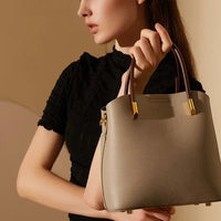 Woman in black top holding taupe leather handbag with brown handles and gold accents