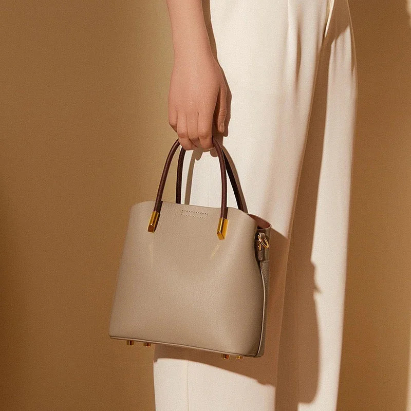 Beige leather handbag with brown handles and gold accents, held by a woman in cream pants