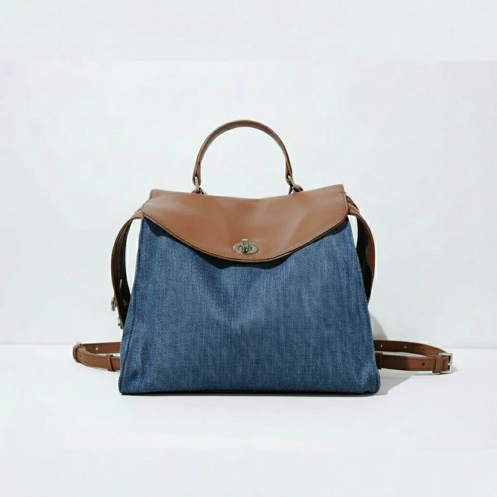 Denim backpack with brown leather flap, handle, and straps on white background