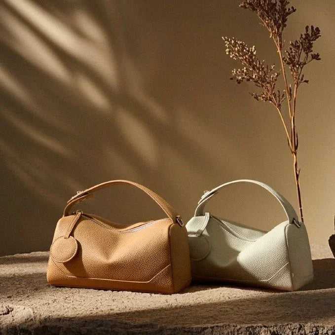 Two leather handbags, tan and light green, displayed on textured stone with dried branch decor