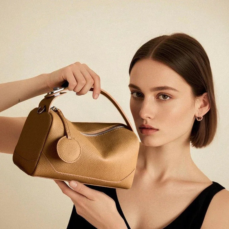 Woman holding tan leather handbag with top handle and round tag against neutral background