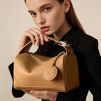 Woman in black outfit holding a tan leather handbag with top handle and round tag