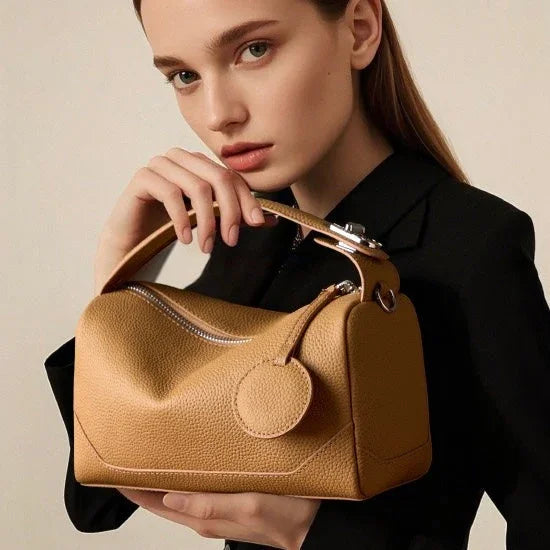 Woman in black outfit holding a tan leather handbag with top handle and round tag