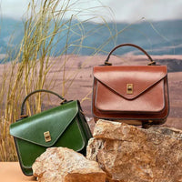 Green and brown leather handbags with top handles on rocks in a nature-inspired outdoor setting