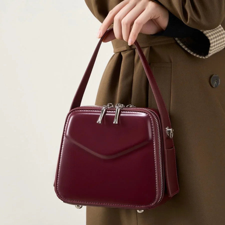 Woman holding a maroon leather handbag with silver zippers, wearing a brown coat