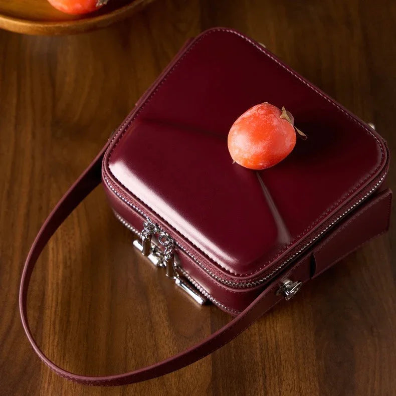 Glossy burgundy square handbag with a persimmon on top, on wooden table
