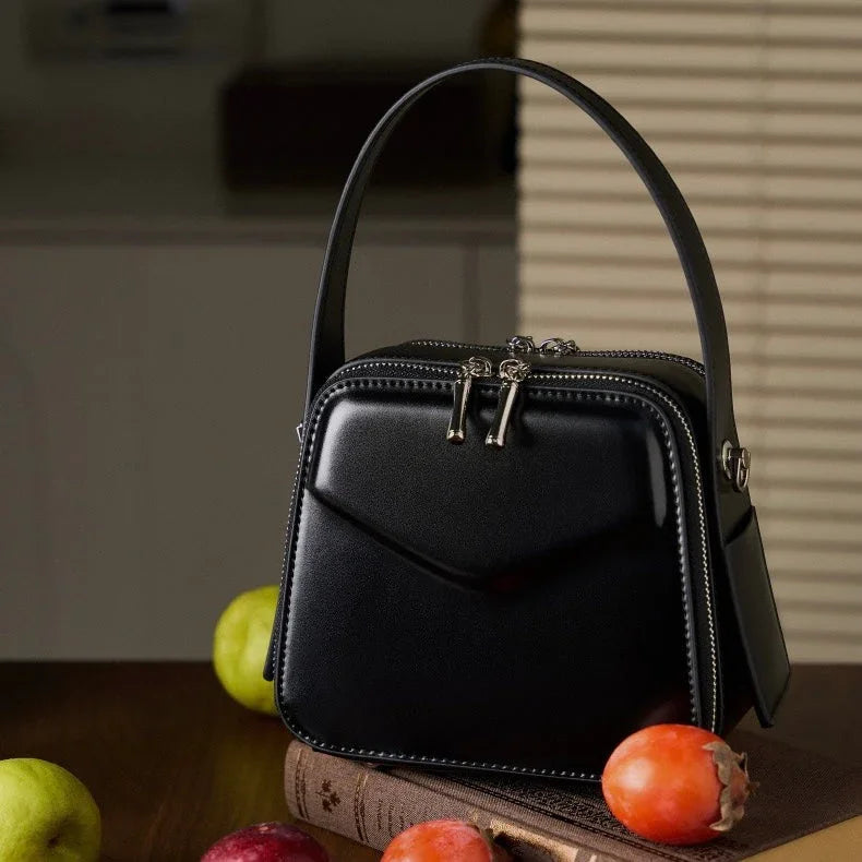 Black leather handbag with top handle on table, fruits and book in background
