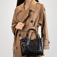Woman in tan trench coat holding a black woven leather handbag against a plain background