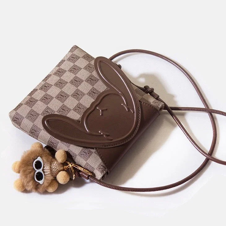 Brown crossbody bag with bunny ear flap and plush bear keychain on checkered fabric