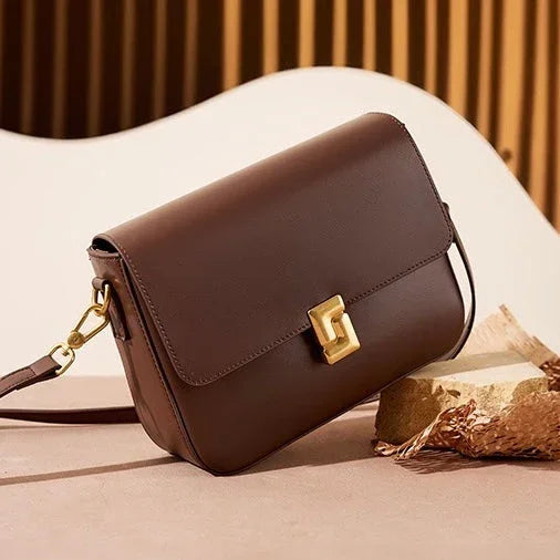 Brown leather crossbody bag with gold clasp on beige surface, modern minimalist setting