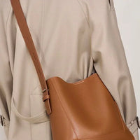 Tan leather bucket bag with adjustable strap worn over a beige trench coat