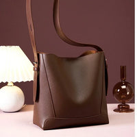 Brown leather bucket bag with wide strap on table, lamp and vase in background