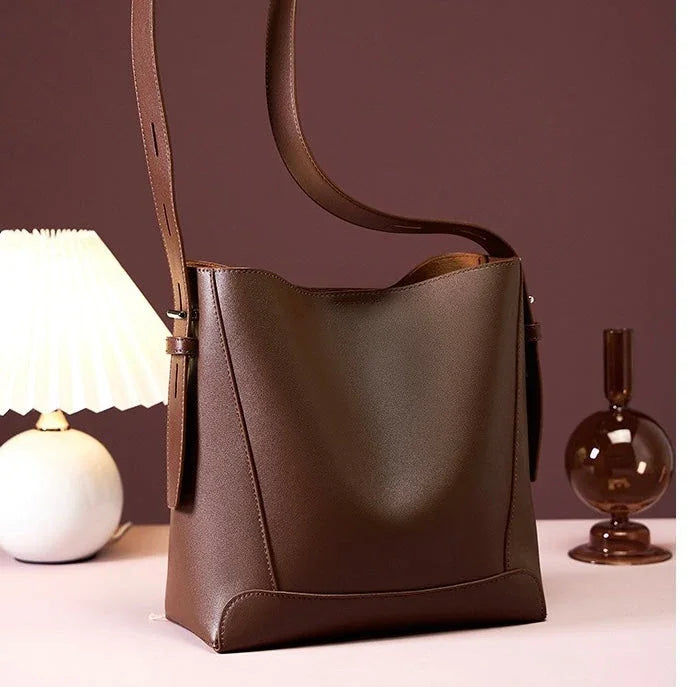 Brown leather bucket bag with wide strap on table, lamp and vase in background