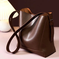 Brown leather tote bag with long strap on table beside white lamp