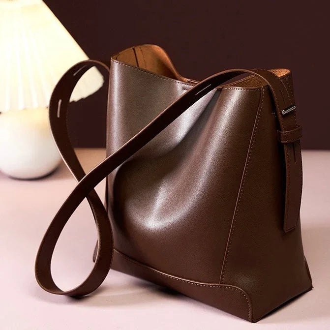 Brown leather tote bag with long strap on table beside white lamp