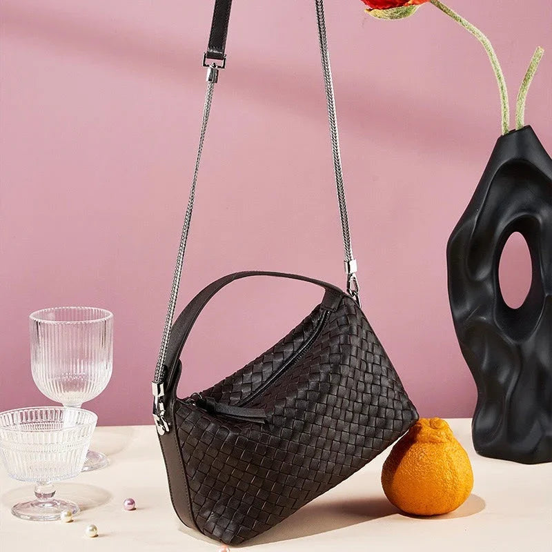 Black woven crossbody bag displayed with glassware, orange, and black vase on pink background