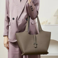 Woman in mauve suit holding large taupe leather tote bag in minimalist indoor setting