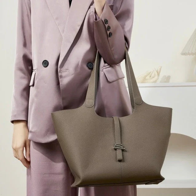 Woman in mauve suit holding large taupe leather tote bag in minimalist indoor setting