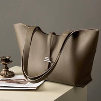 Taupe leather tote bag with double straps on a table beside a brass candle holder and magazine