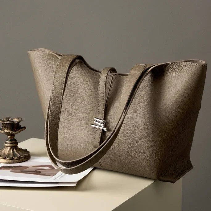 Taupe leather tote bag with double straps on a table beside a brass candle holder and magazine