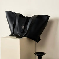 Black leather tote bag with long straps on a beige pedestal, minimalist setting