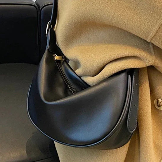 Black leather crescent shoulder bag on beige coat, modern minimalist fashion accessory