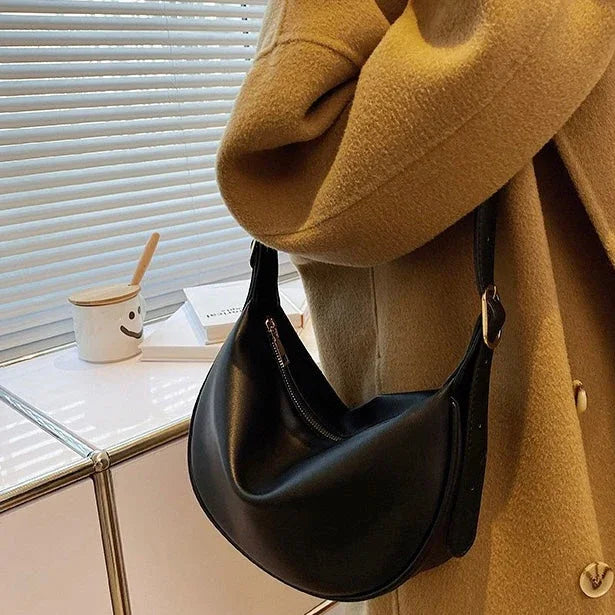 Woman in camel coat holding black leather shoulder bag indoors by window blinds