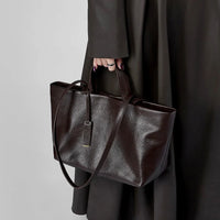 Hand holding a dark brown leather tote bag with tag, black coat in background