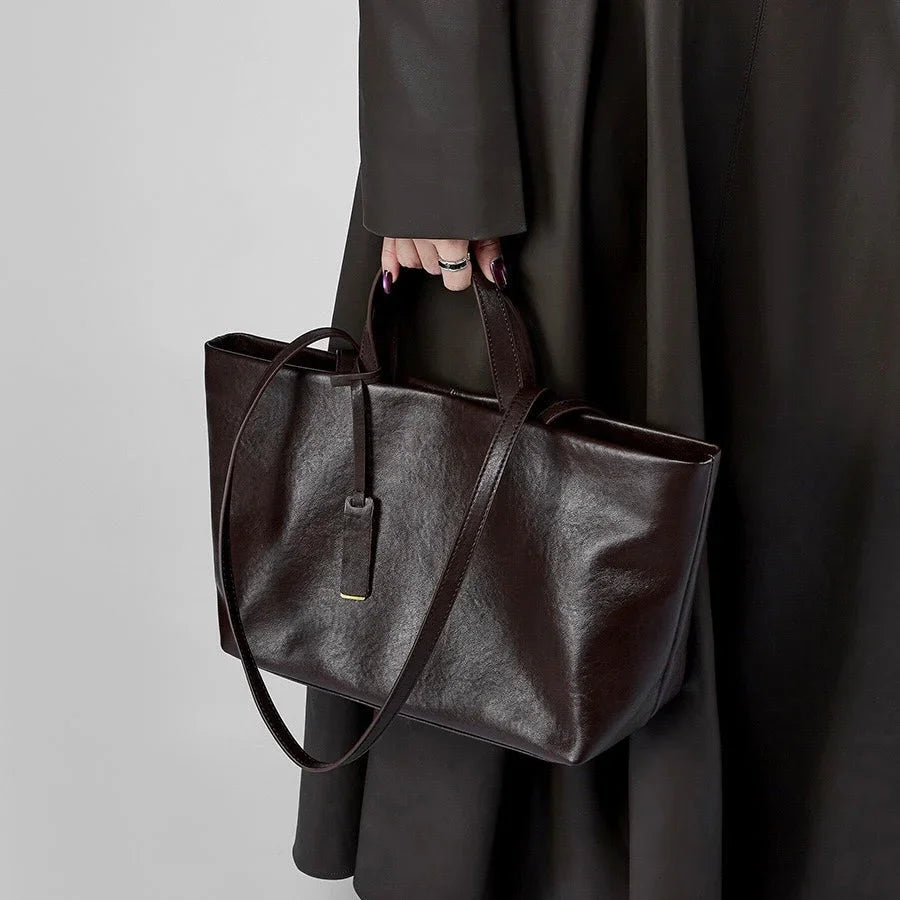 Hand holding a dark brown leather tote bag with tag, black coat in background