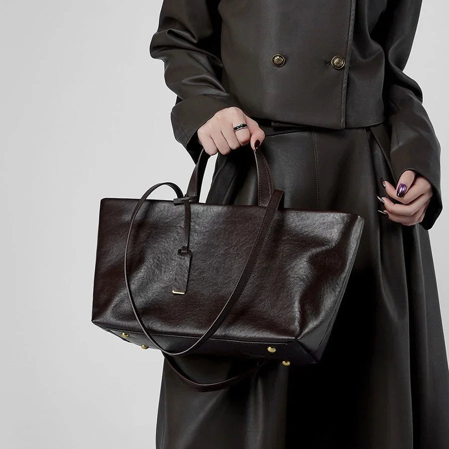 Woman in dark leather outfit holding a large brown leather tote bag with gold accents