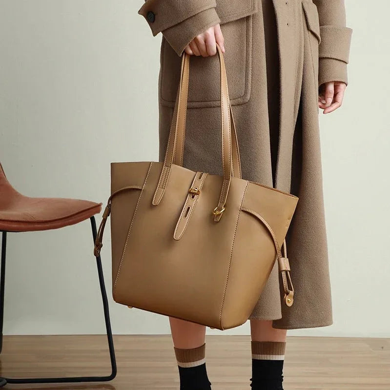 Woman in tan coat holding large brown leather tote bag indoors next to modern chair