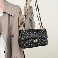 Woman in beige trench coat holding black quilted leather shoulder bag with gold chain strap