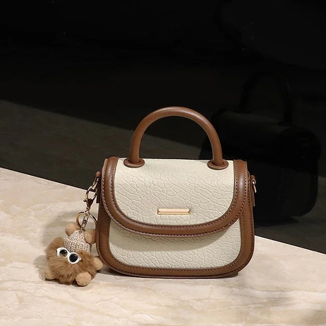 Beige and brown textured leather handbag with top handle and plush bear keychain on marble surface