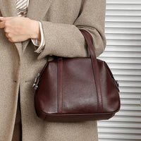 Woman in beige coat holding a brown leather handbag in front of a striped background