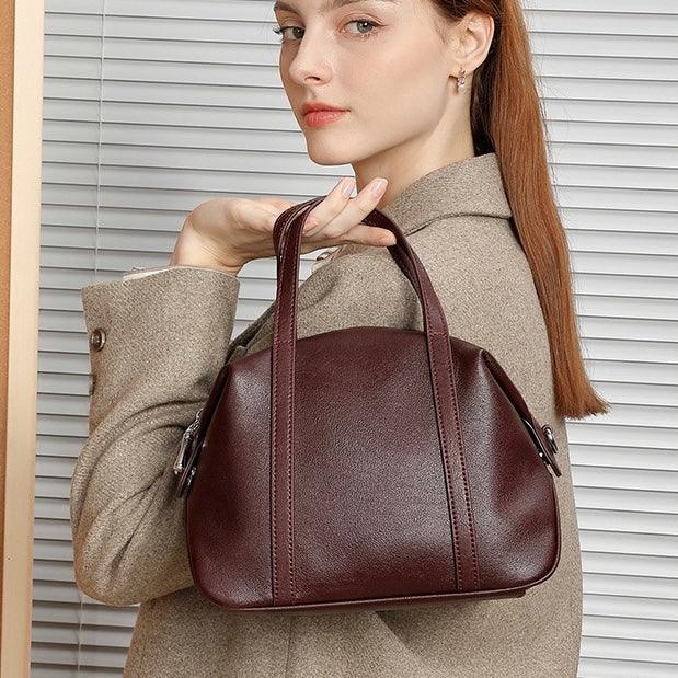 Woman in beige coat holding a brown leather handbag in front of blinds