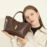 Woman holding a stylish brown leather handbag with gold clasp, cream jacket, minimalist background