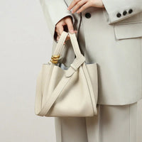Woman in a cream suit holding a beige leather handbag with gold accent and shoulder strap
