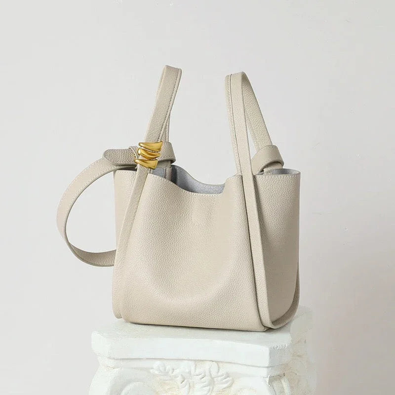 Beige leather tote bag with gold accent on strap, minimalist style, on decorative pedestal