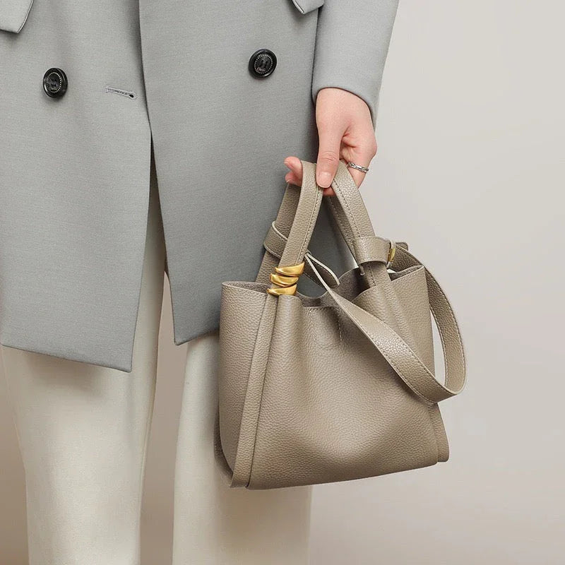 Woman holding taupe leather bucket bag, wearing light grey coat and white pants