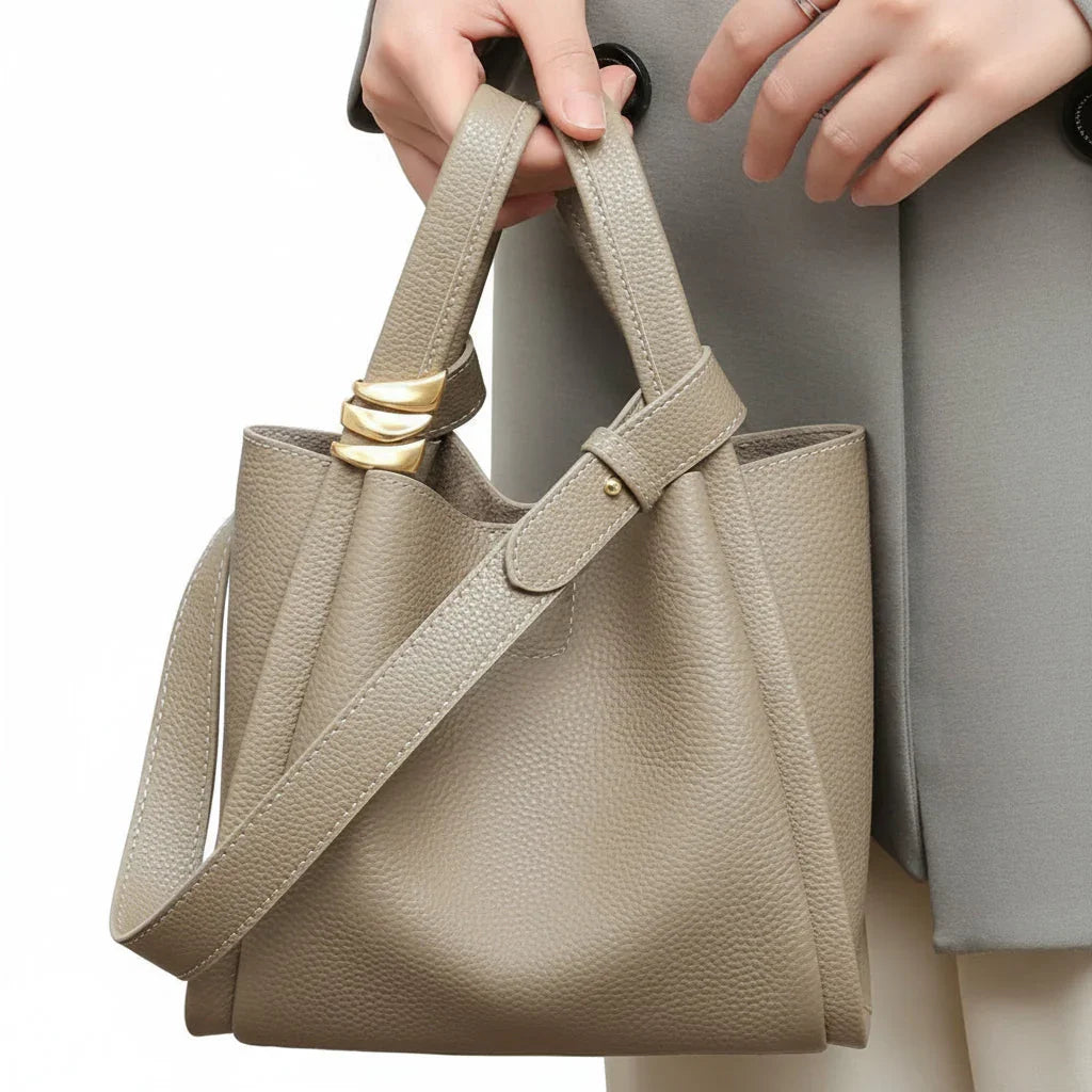 Taupe leather bucket bag with gold accents and shoulder strap held by woman in grey coat