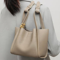 Taupe leather tote bag with gold rings on handle, worn over a grey blazer