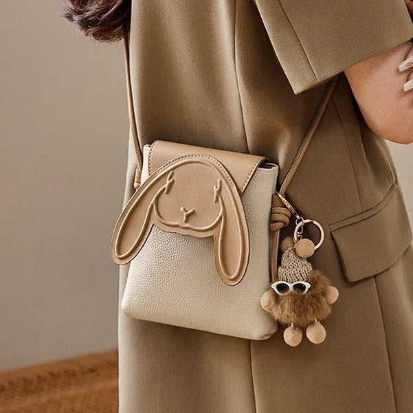 Beige crossbody bag with bunny face design and cute plush keychain, worn on shoulder.