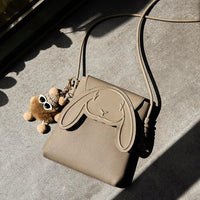 Beige crossbody bag with bunny face design and plush keychain on a concrete surface