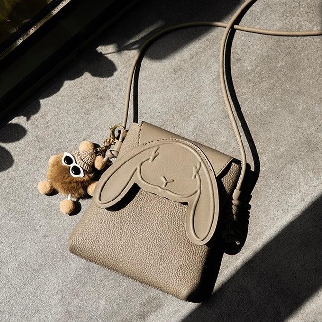 Beige crossbody bag with bunny face design and plush keychain on a concrete surface