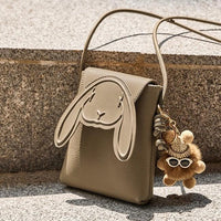 Beige bunny crossbody bag with plush bear keychain, outdoor stone background