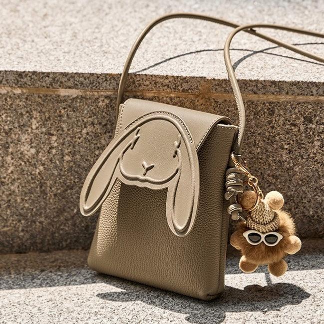 Beige bunny crossbody bag with plush bear keychain, outdoor stone background